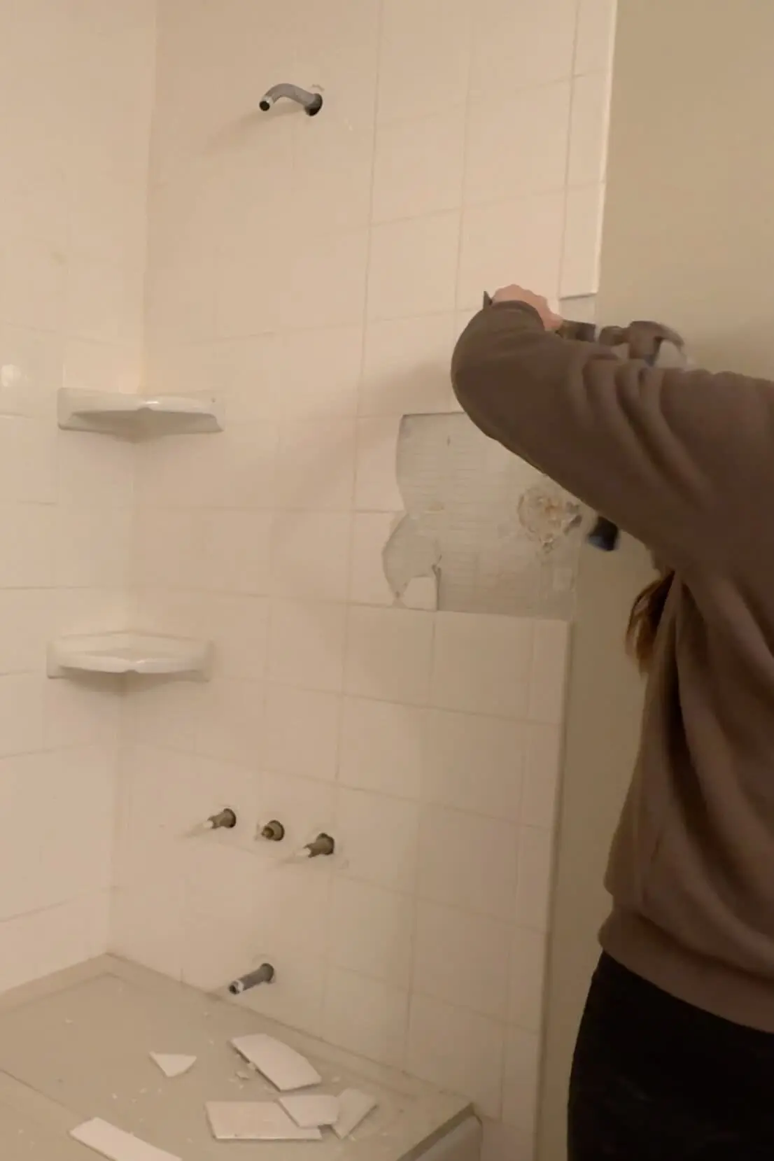 A person wearing safety goggles, a brown sweatshirt, and black leggings is shown using a demolition tool to remove white tiles from a bathroom wall. Broken tiles and debris scatter on a countertop below as the remodel begins.
